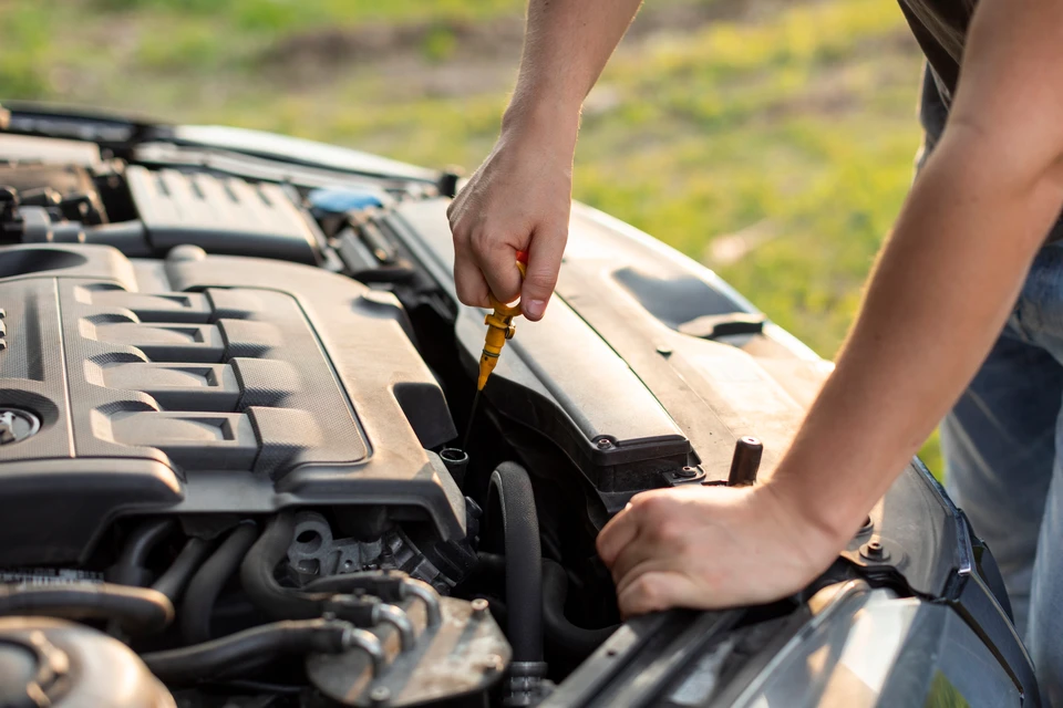 Mécanicien automobile inspecte un moteur sous le capot ouvert d'un véhicule, tenant un carnet de maintenance avec grille de données et stylo pour noter les vérifications techniques